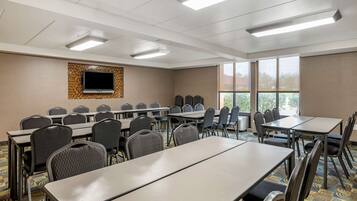 A conference room with multiple tables and chairs, a flat-screen TV mounted on the wall, and large windows providing natural light.