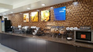 Zona de bufé con salpicadero de azulejos, una pantalla grande que muestra 'RISE & SHINE' y diferentes utensilios para servir comida.