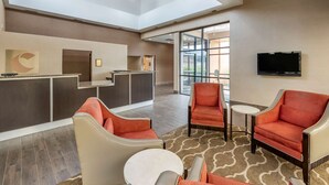 Un vestíbulo de hotel con una recepción, un área de estar con sillas y una mesa, un televisor montado en la pared y un gran ventanal que proporciona luz natural.