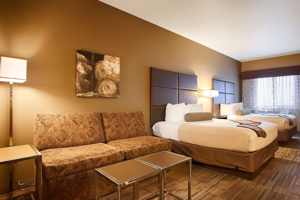 A hotel room with a sofa, two beds, a small table, and a painting on the wall.