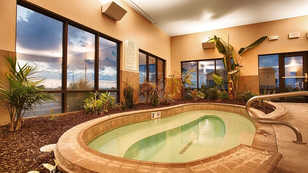 Una piscina interior con un cartel que indica una profundidad de 3 pies, rodeada de plantas y grandes ventanales que ofrecen vistas al cielo y edificios.