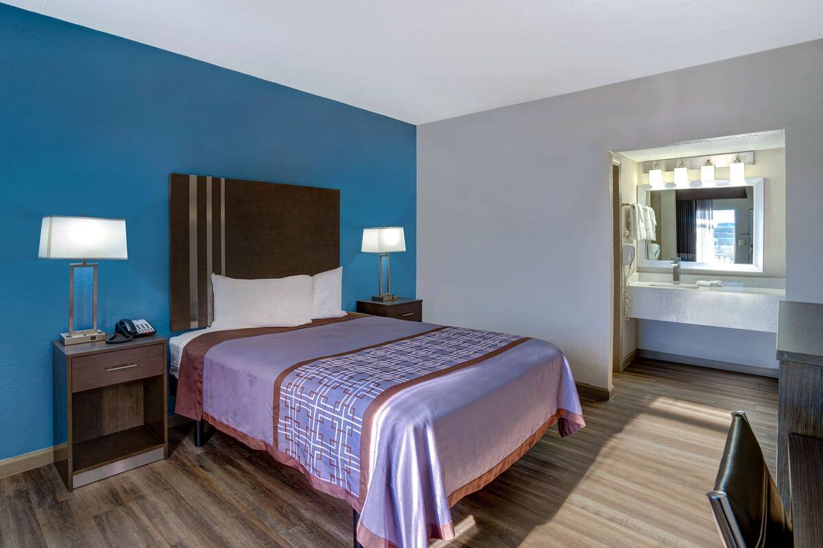 Habitación de hotel con una cama grande, dos mesitas de noche con lámparas, una mesita auxiliar, un baño con espejo y lavamanos, y suelo de madera.