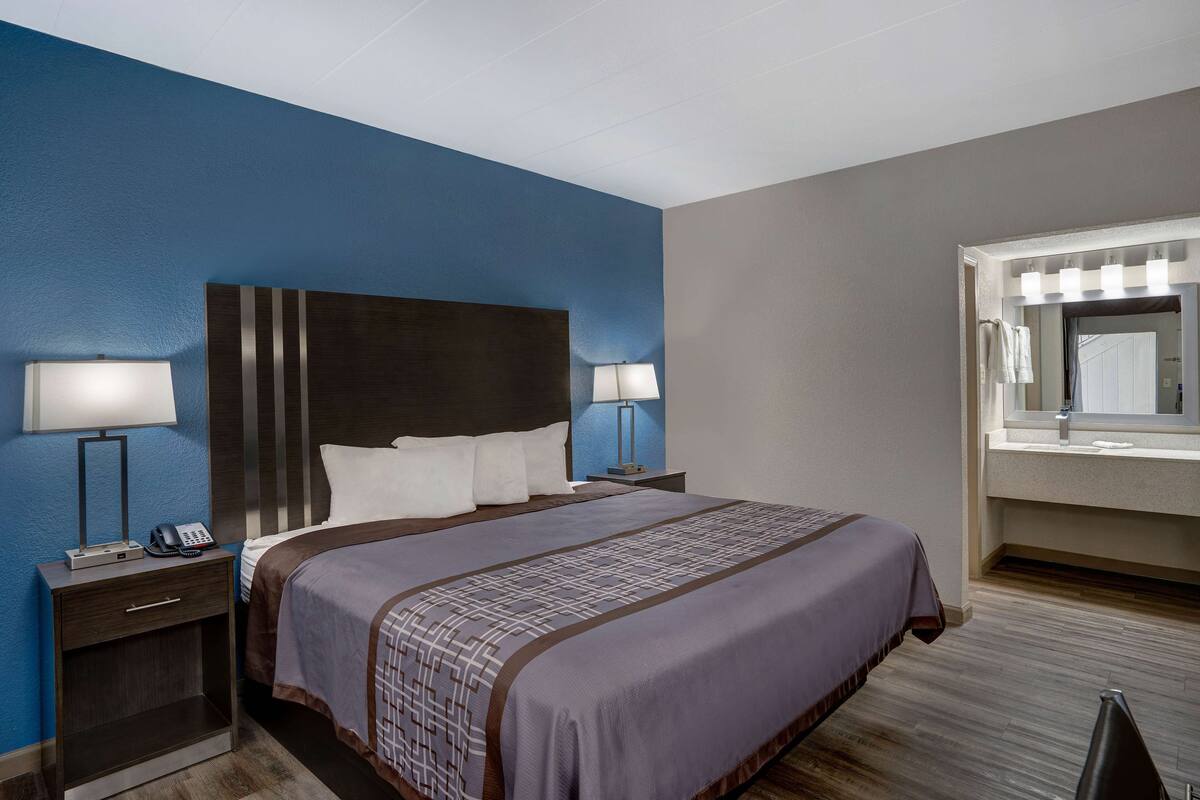 Habitación de hotel con una cama grande, dos mesitas de noche con lámparas, un espejo sobre el lavabo y una pared azul de acento.