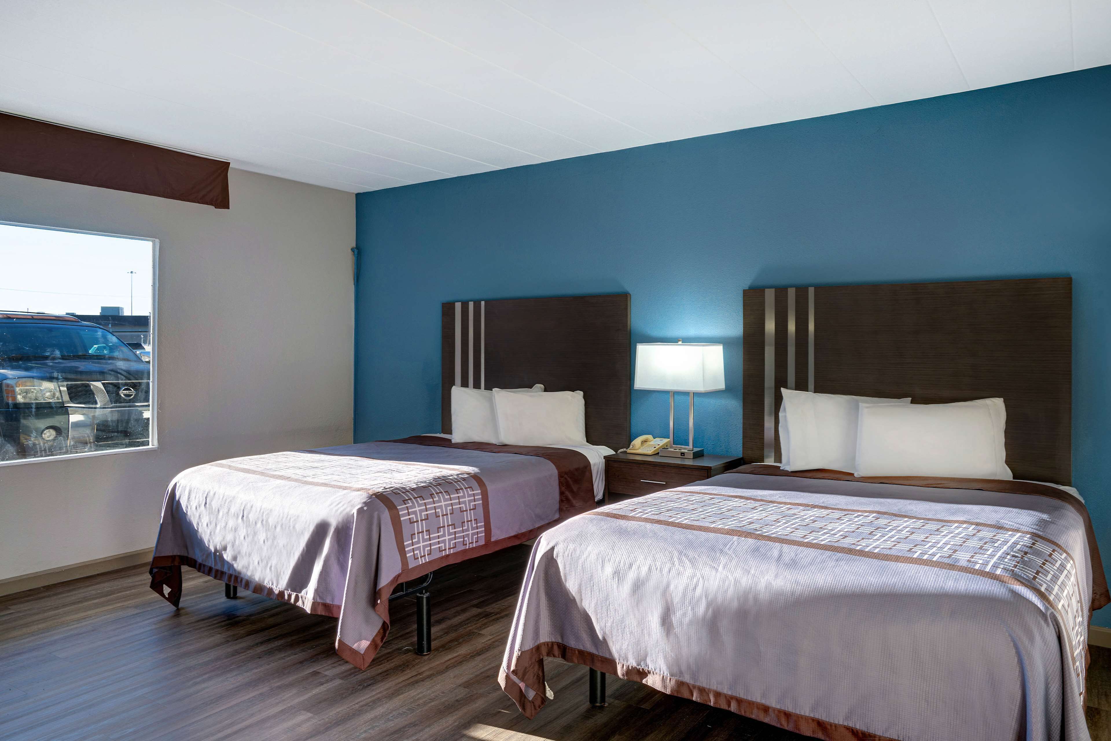 A hotel room with two beds, a window with a view of parked cars, and a blue wall.
