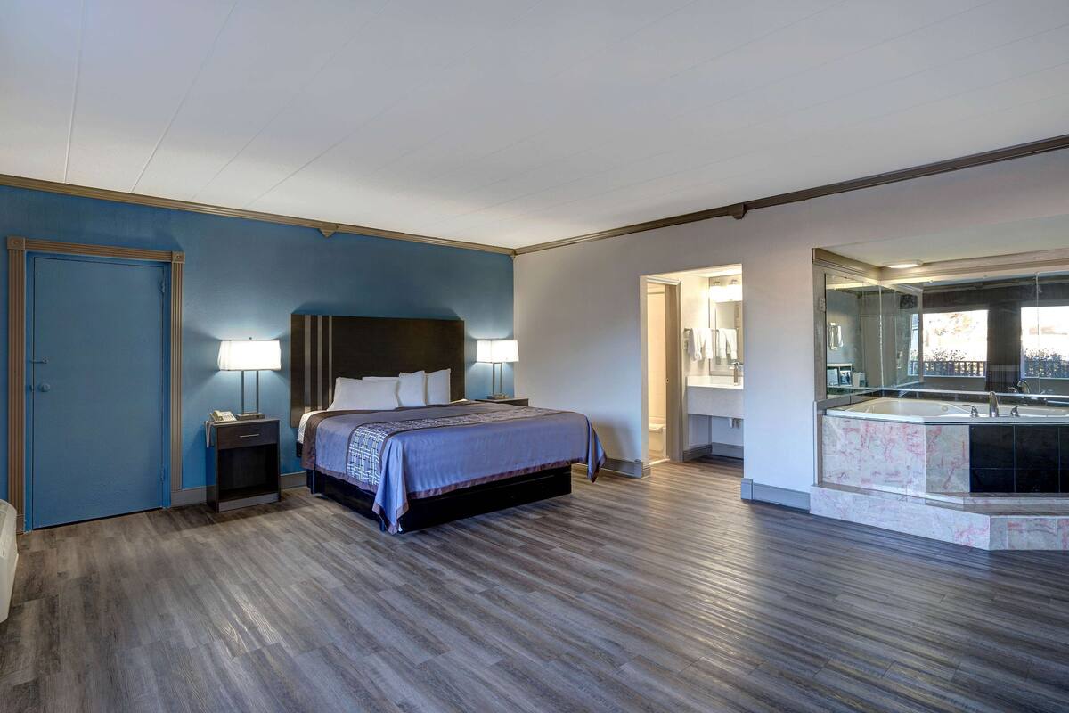 Una habitación de hotel moderna con una cama grande, lámparas de noche, un escritorio integrado y un baño con un espejo grande y lavabo.
