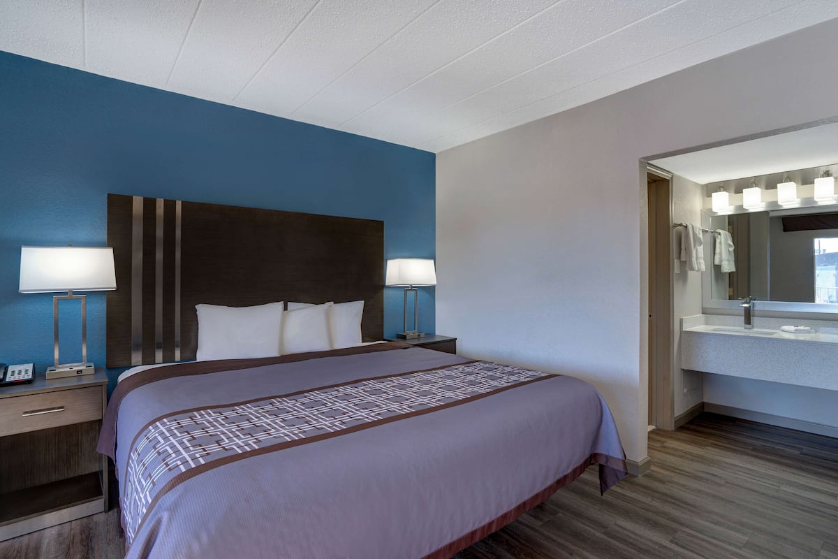 Una habitación de hotel con una cama grande, lámparas de noche, una mesita de noche y un baño con lavamanos y espejo.