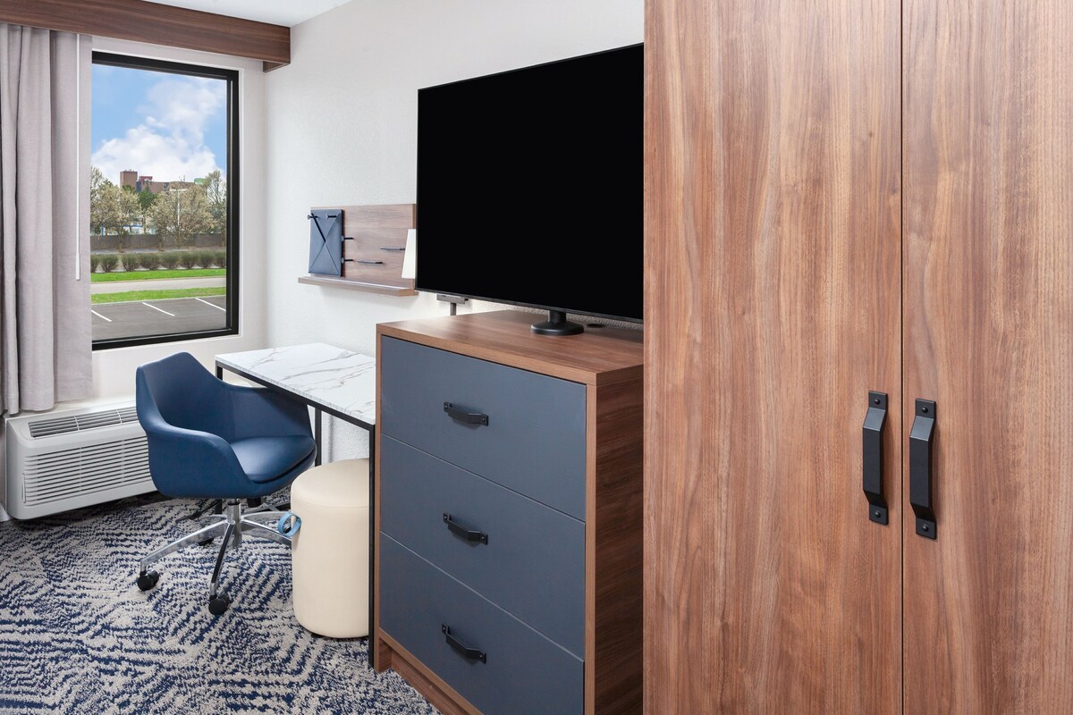 Une chambre d’hôtel avec une grande fenêtre, un bureau, une chaise, une télévision et une armoire.