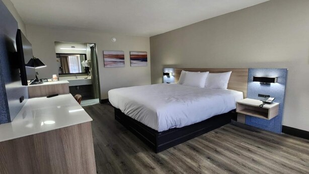A modern hotel room with a large bed, a desk with a chair, a vanity with a mirror, and two wall-mounted lamps. - Baymont by Wyndham Lafayette (Lafayette)