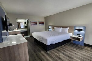 A modern hotel room with a large bed, a desk with a chair, a vanity with a mirror, and two wall-mounted lamps.