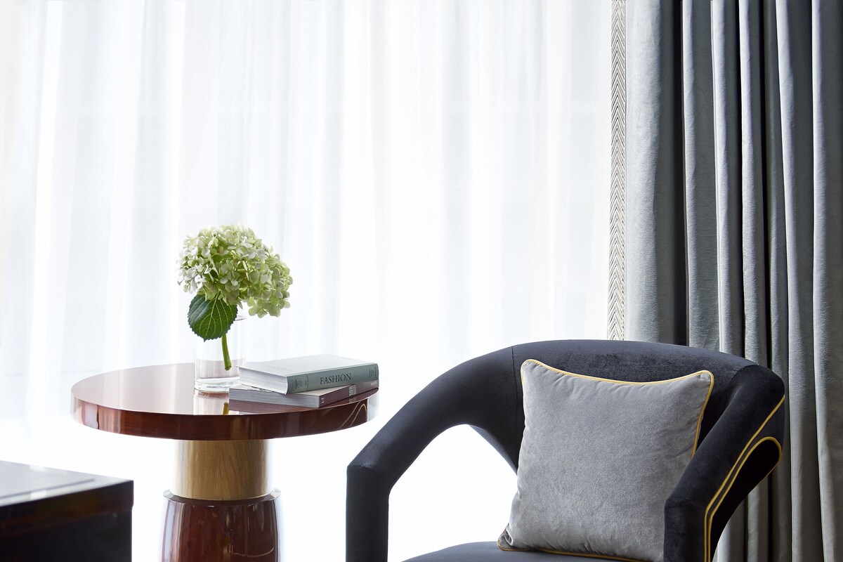 A modern living room with a dark upholstered armchair, a round wooden side table with a vase of flowers, and a stack of books.