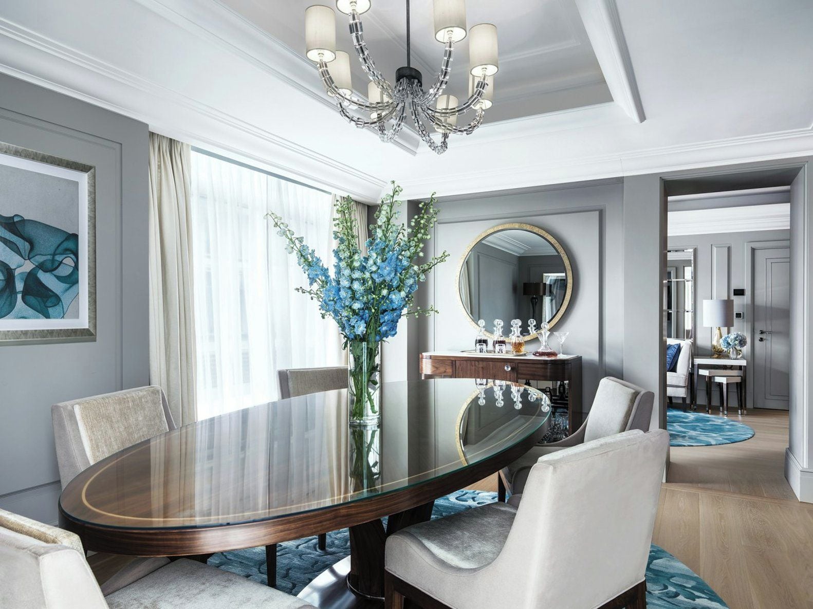 A modern dining area with a glass-top table, white upholstered chairs, a vase of blue flowers, a mirror above the table, and a chandelier.