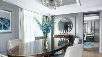 A modern dining area with a glass-top table, white upholstered chairs, a vase of blue flowers, a mirror above the table, and a chandelier.