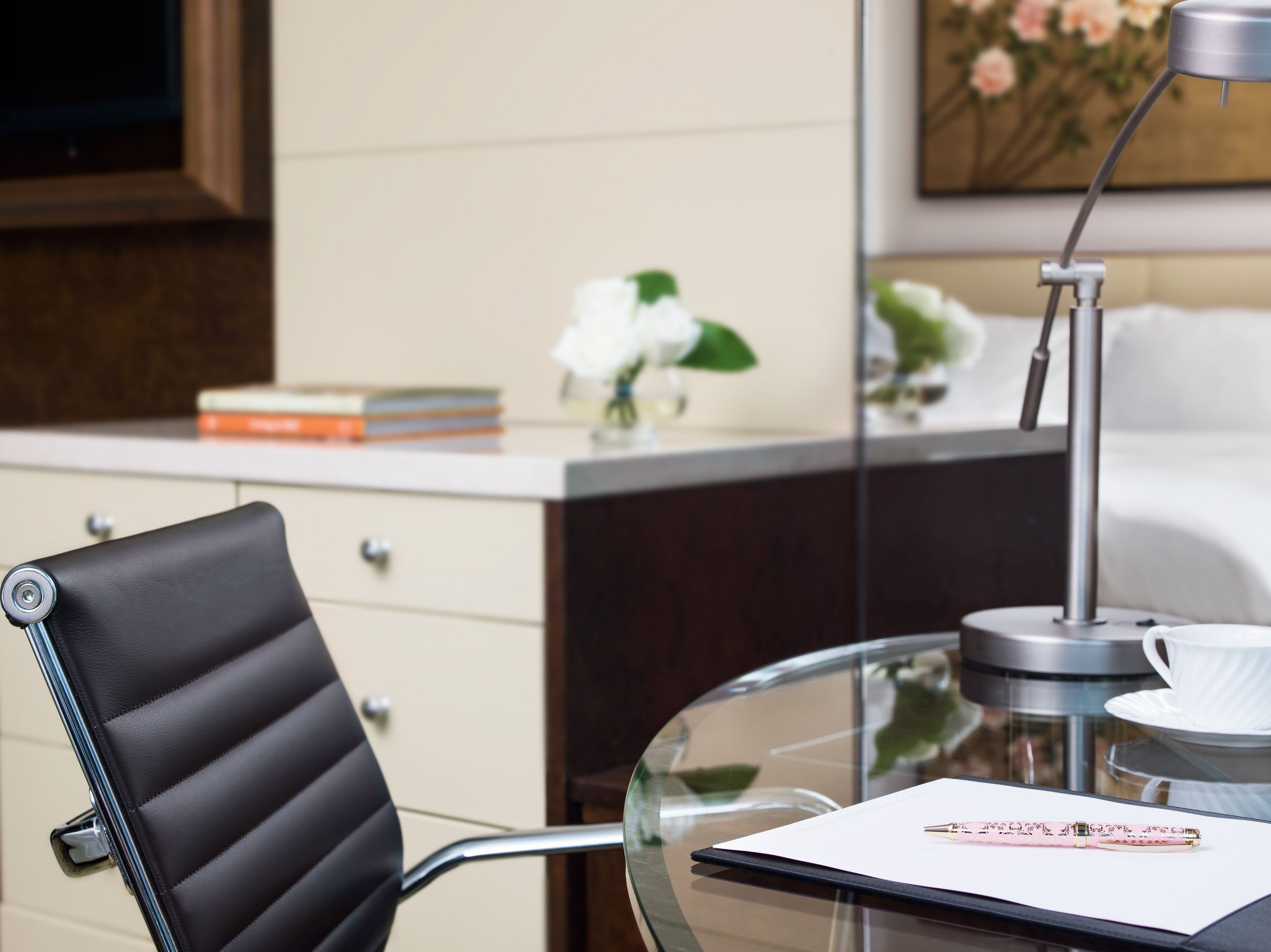 A modern hotel room with a glass table, a black leather chair, a bedside table with a lamp, and a bed with white bedding.