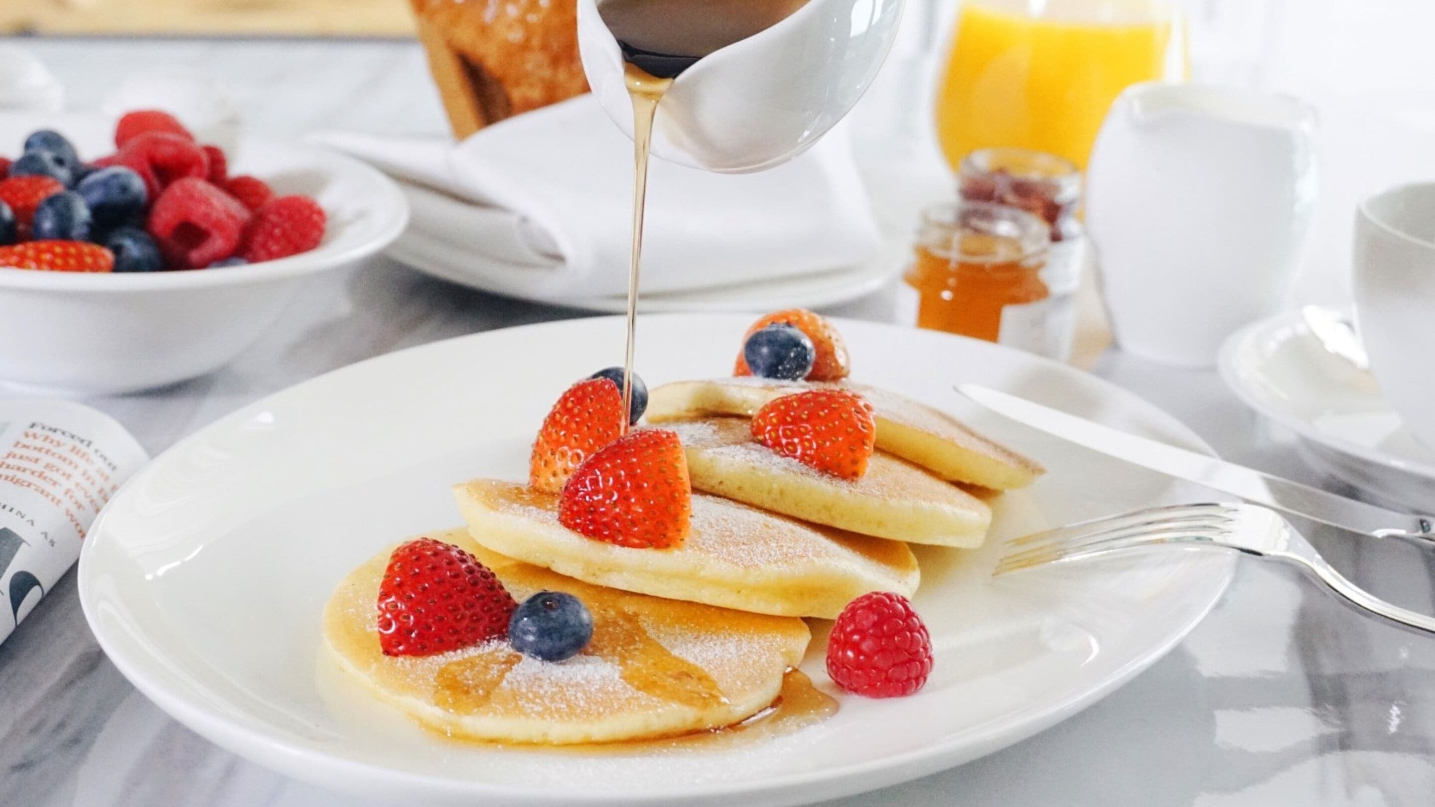 A breakfast plate with pancakes topped with strawberries and blueberries, syrup being poured, and a bowl of mixed berries in the background.