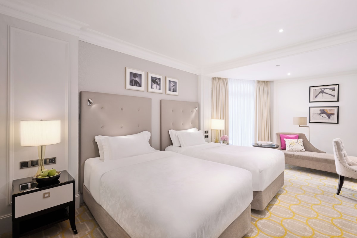 A hotel room with two beds, a nightstand with a lamp, a chair, and a small table with a bowl of fruit.