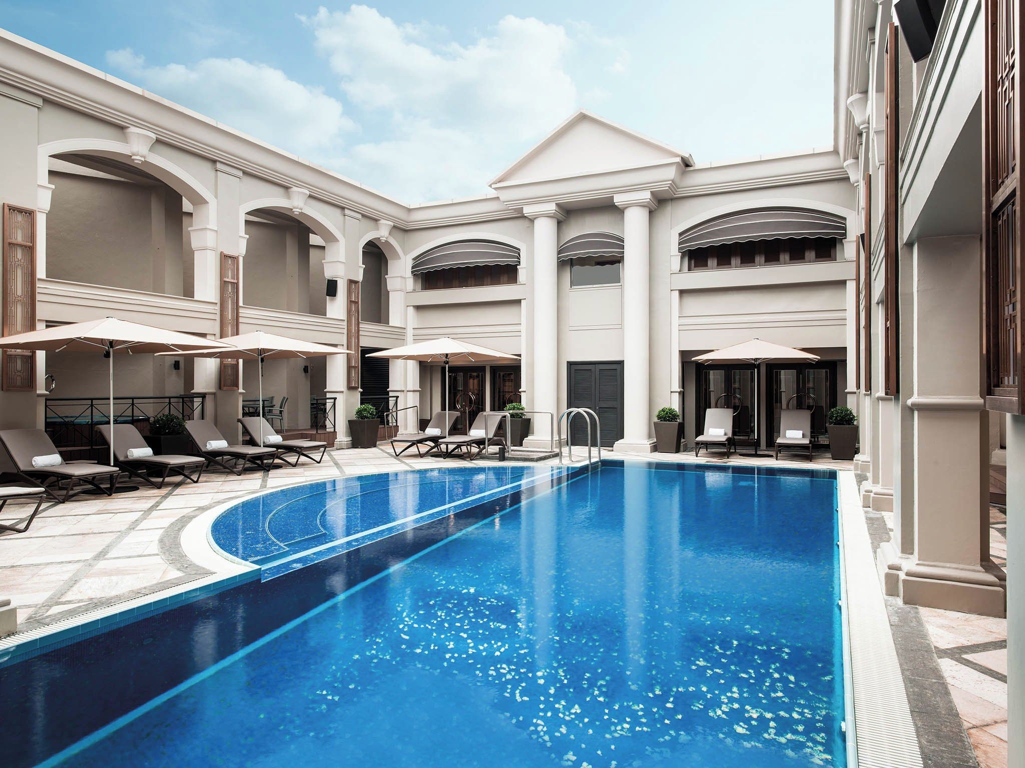 A swimming pool with lounge chairs and umbrellas, surrounded by a building with arches and columns.