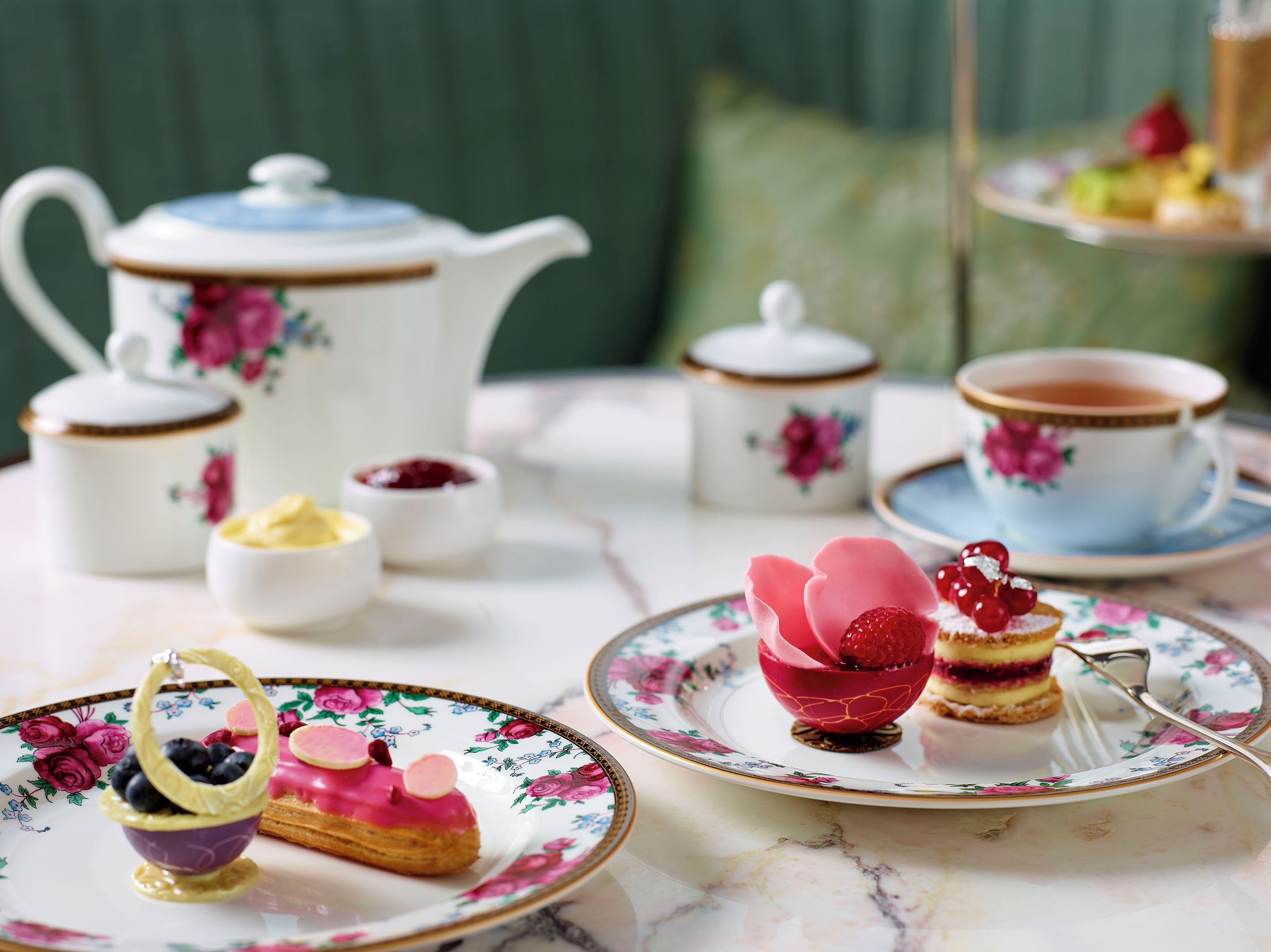 A tea setting with floral-patterned tableware, including a teapot, cups, and plates, with a dessert on a floral plate.
