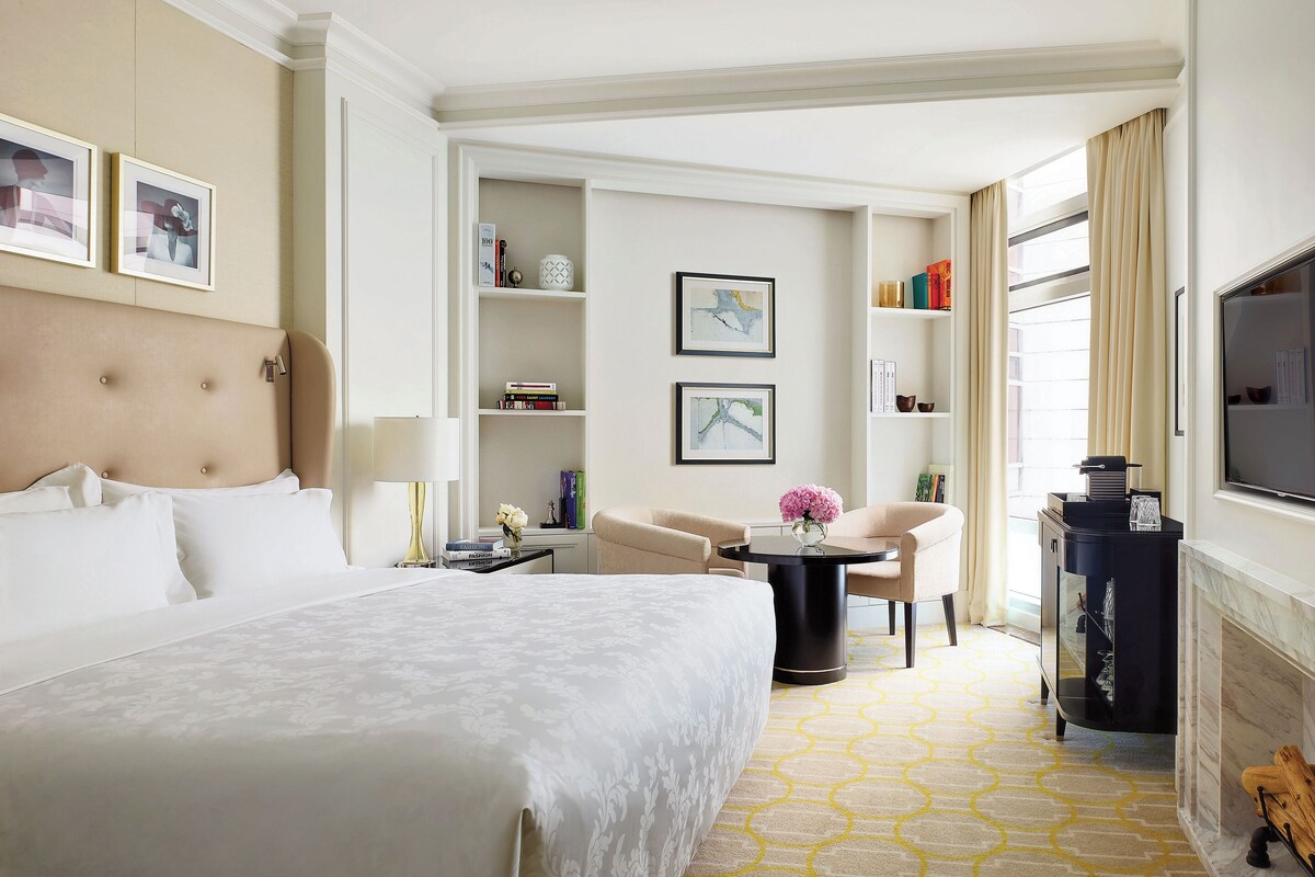 A hotel room with a large bed, a desk, a chair, a TV, and a fireplace.