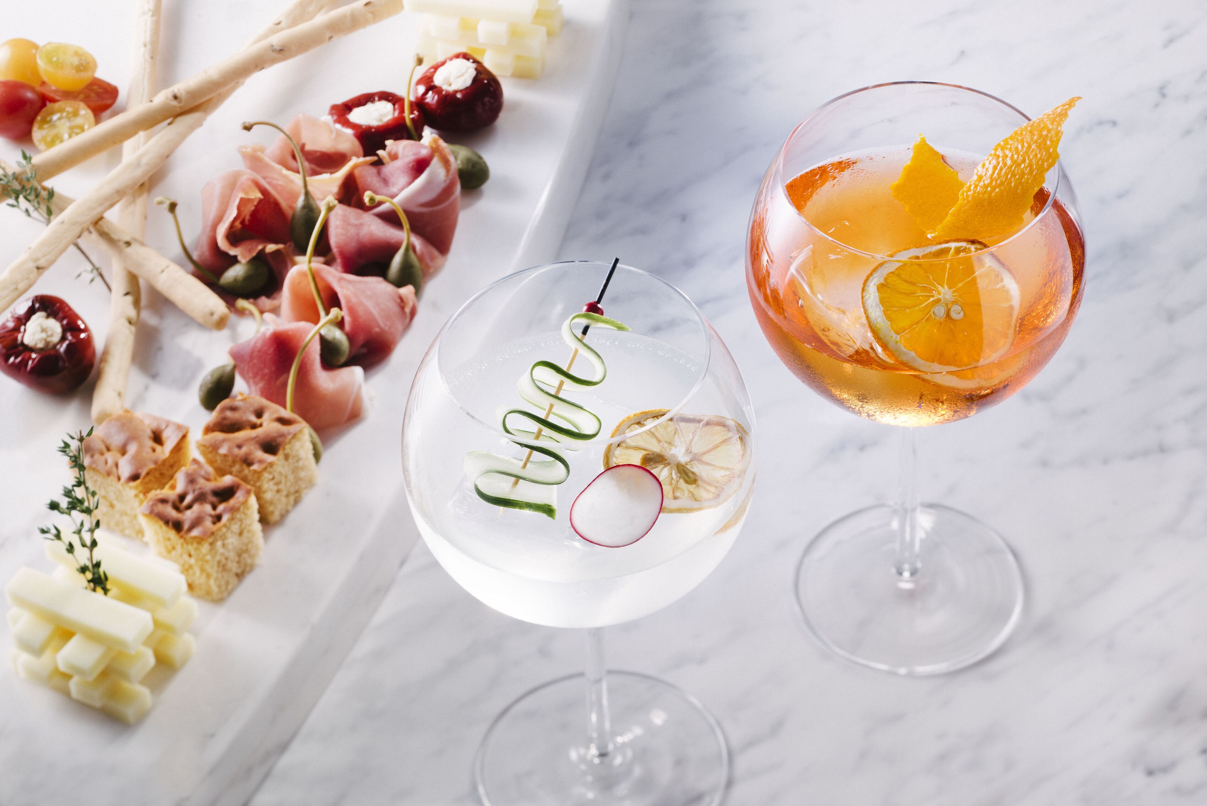 Dos vasos de cócteles con adornos, una variedad de aperitivos que incluyen jamón serrano y queso, y una selección de frutas y frutos secos sobre una superficie de mármol.