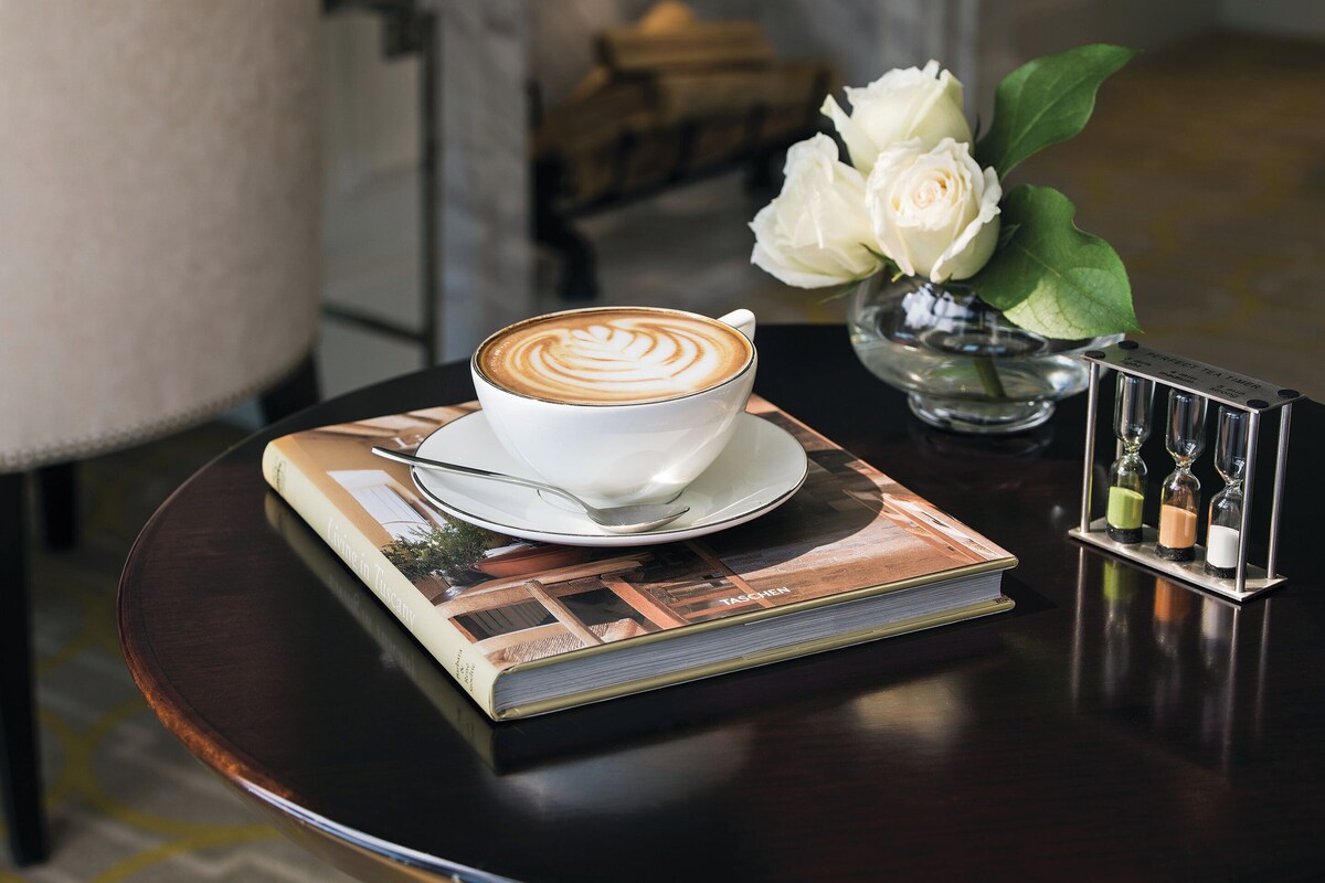 A cup of coffee with latte art on a saucer, a book titled "In Tuscany," a vase with white flowers, and a small hourglass display on a table.