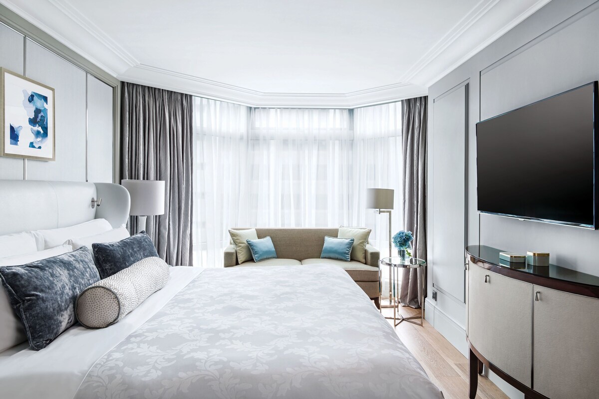 A modern hotel room with a large bed, a flat-screen TV, a seating area with a sofa, and a dining table.