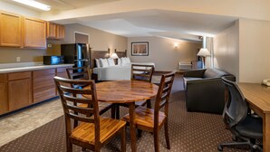 A hotel room with a kitchenette, a dining area with a wooden table and chairs, a bed with a white headboard, and a desk with a chair. - Best Western Desert Inn (West Yellowstone)