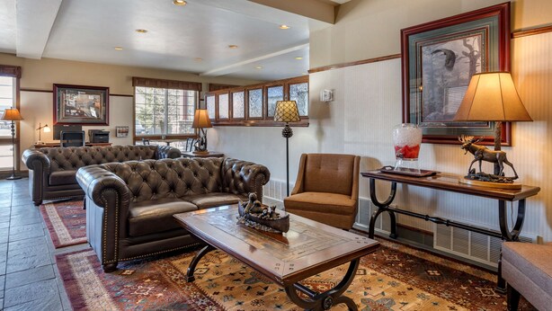 A spacious living room with a large leather sofa, a smaller armchair, and a coffee table. There are multiple windows providing natural light, and a framed picture on the wall. - Best Western Desert Inn (West Yellowstone)