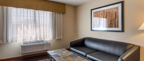 A hotel room with a black leather sofa, a patterned coffee table, a large window with sheer curtains, a framed picture on the wall, and a standing heater.