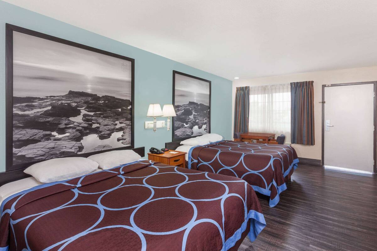 A hotel room with two beds, a nightstand, a lamp, and a large framed photograph on the wall.