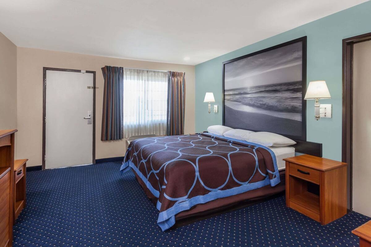 A hotel room with a bed, a nightstand, a lamp, a painting on the wall, and a door.