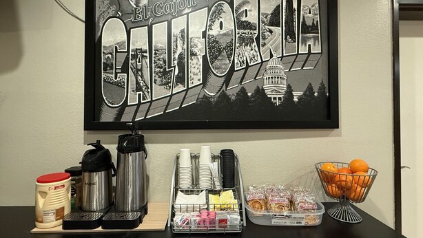 Un letrero en la pared con la inscripción 'Greetings from California' y una estación de café con cafetera, tazas y condimentos.