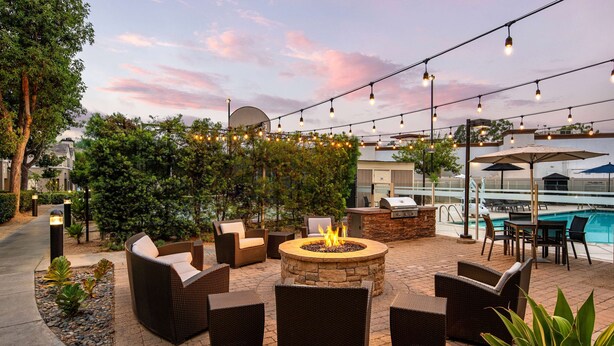 An outdoor patio with wicker furniture, a stone fire pit, string lights, and a pool area. - Sonesta ES Suites Carmel Mountain – San Diego (San Diego)