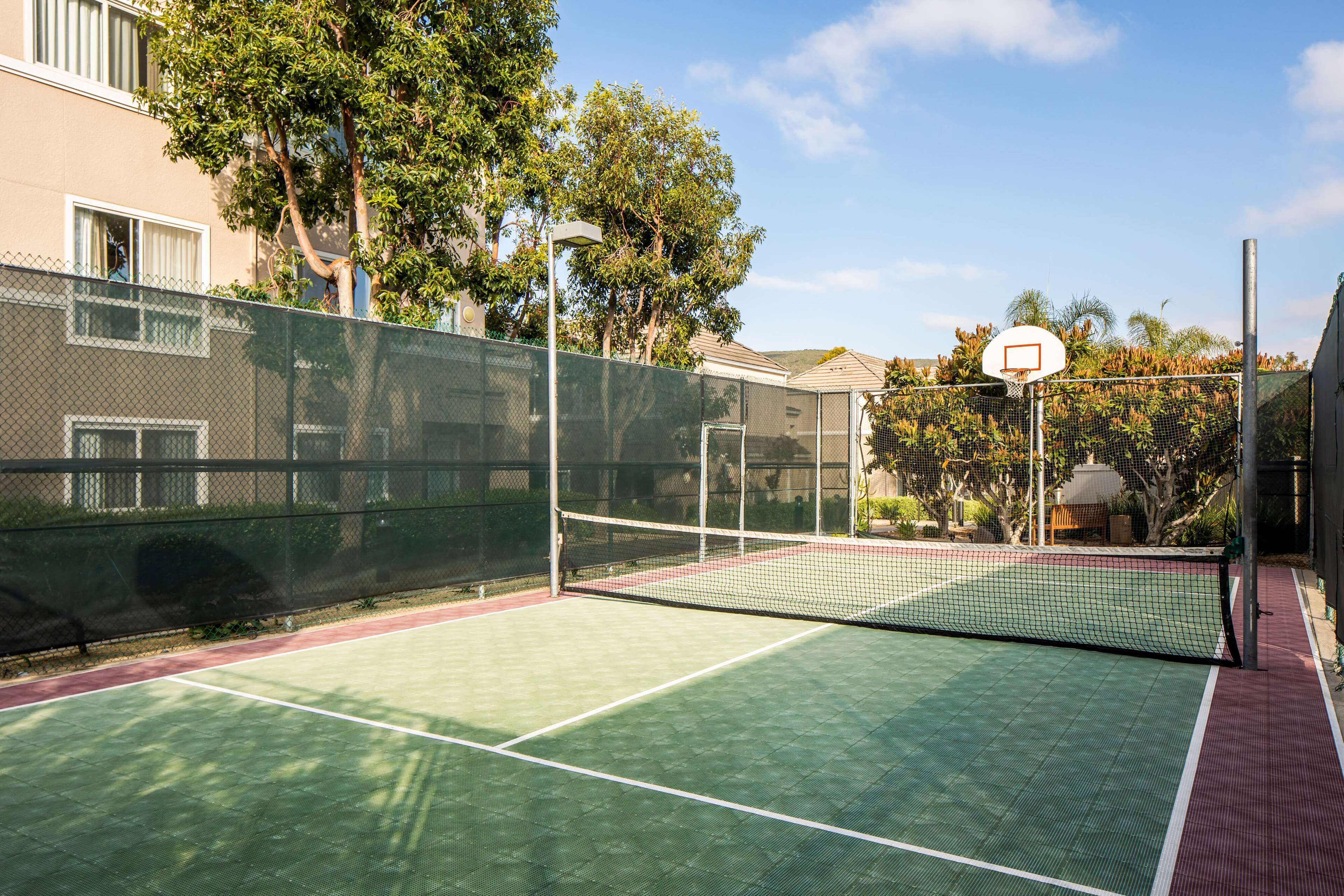 An outdoor tennis court with a net, surrounded by a fence and trees.