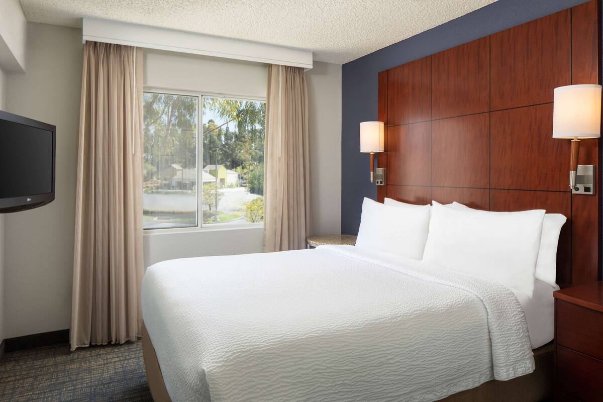 Een hotelkamer met een groot bed, een televisie, een raam met gordijnen en een houten hoofdeinde met wandlampen.