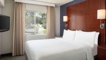 A hotel room with a large bed, a TV, a window with curtains, and a wooden headboard with wall-mounted lamps.