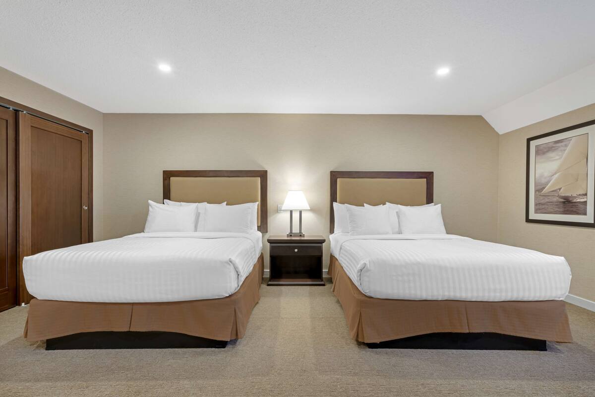 A hotel room with two beds, a nightstand with a lamp, and a large window with a sailboat view.