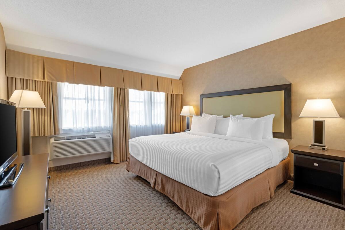 A hotel room with a large bed, two bedside lamps, a flat-screen TV, a desk with a lamp, and a window with curtains.