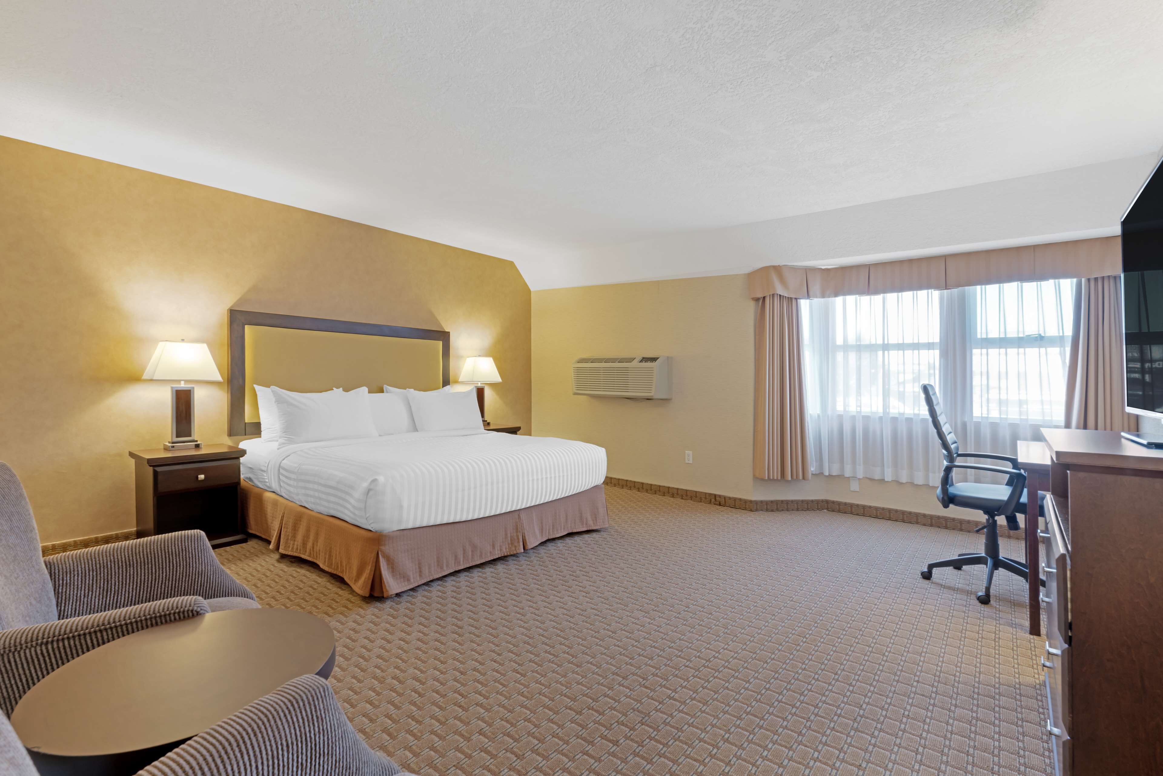 Habitación de hotel con cama, dos mesitas de noche con lámparas, un escritorio con silla, un equipo de aire acondicionado y un ventanal con cortinas.