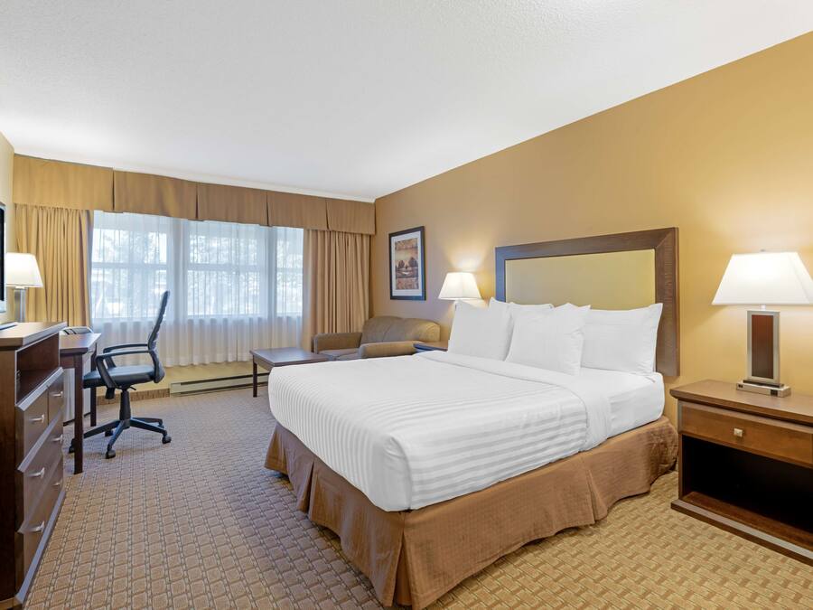 A hotel room with a large bed, a desk with a computer, a chair, a desk lamp, and a bedside table with a lamp.