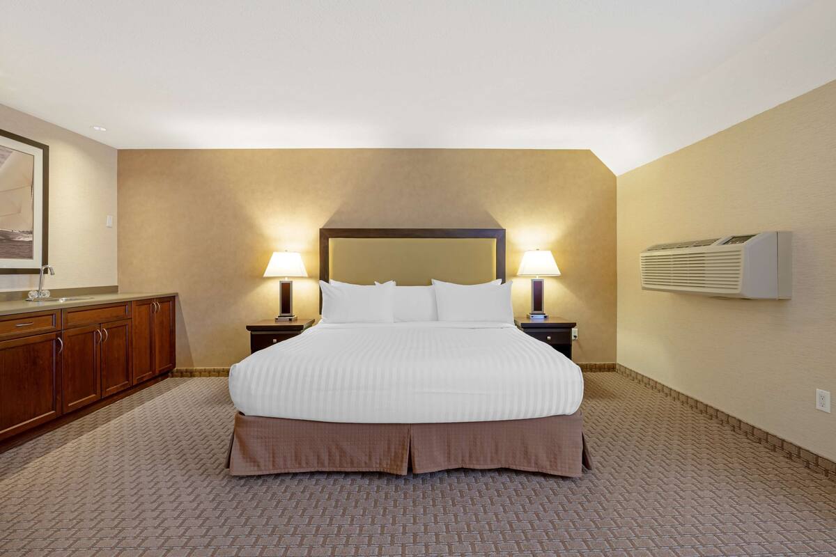 A hotel room with a large bed, two bedside lamps, a wooden dresser, and an air conditioning unit.