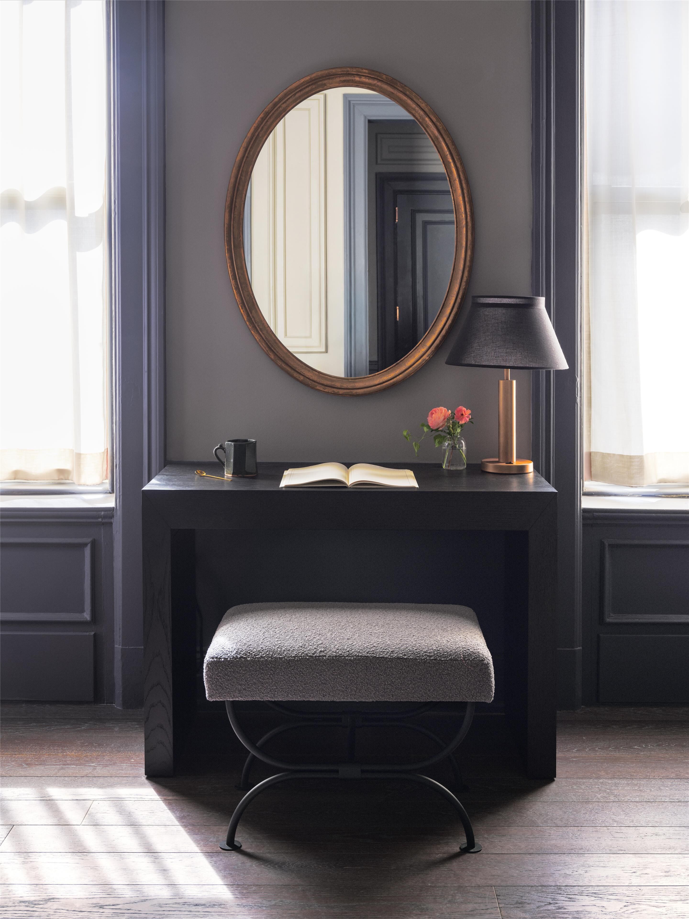 A dark-toned vanity with a round mirror, a small bench, and a lamp.