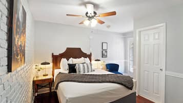 Une chambre à coucher avec un lit, une table de chevet, une lampe, un ventilateur de plafond, une porte et un mur en briques.
