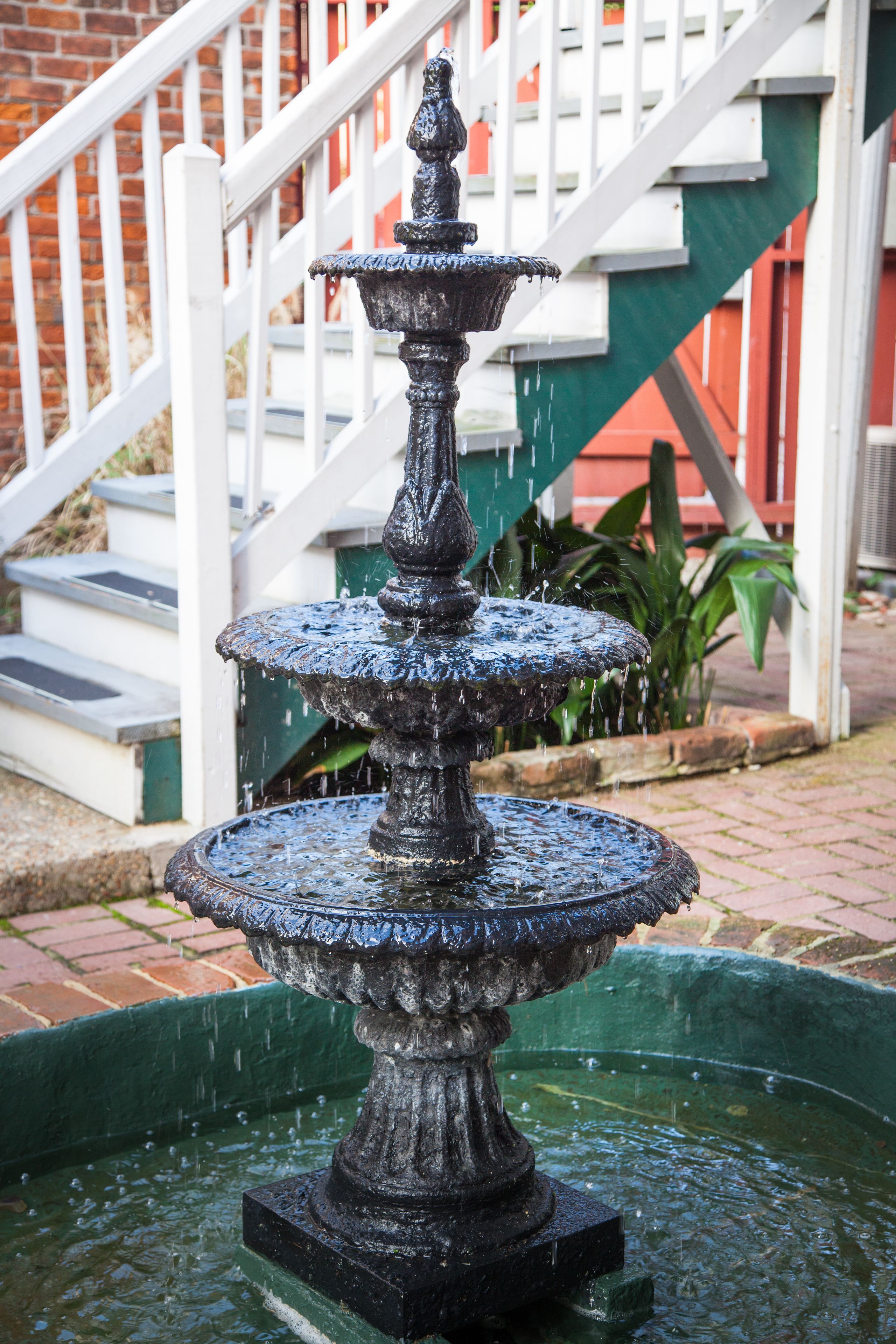 Une fontaine à plusieurs niveaux, dont l’eau jaillit en cascade, est adossée à un mur de briques et agrémentée d’une balustrade blanche.