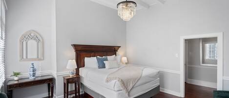 A bedroom with a large bed, a nightstand, a chair, a small table, a chandelier, and a door leading to another room.