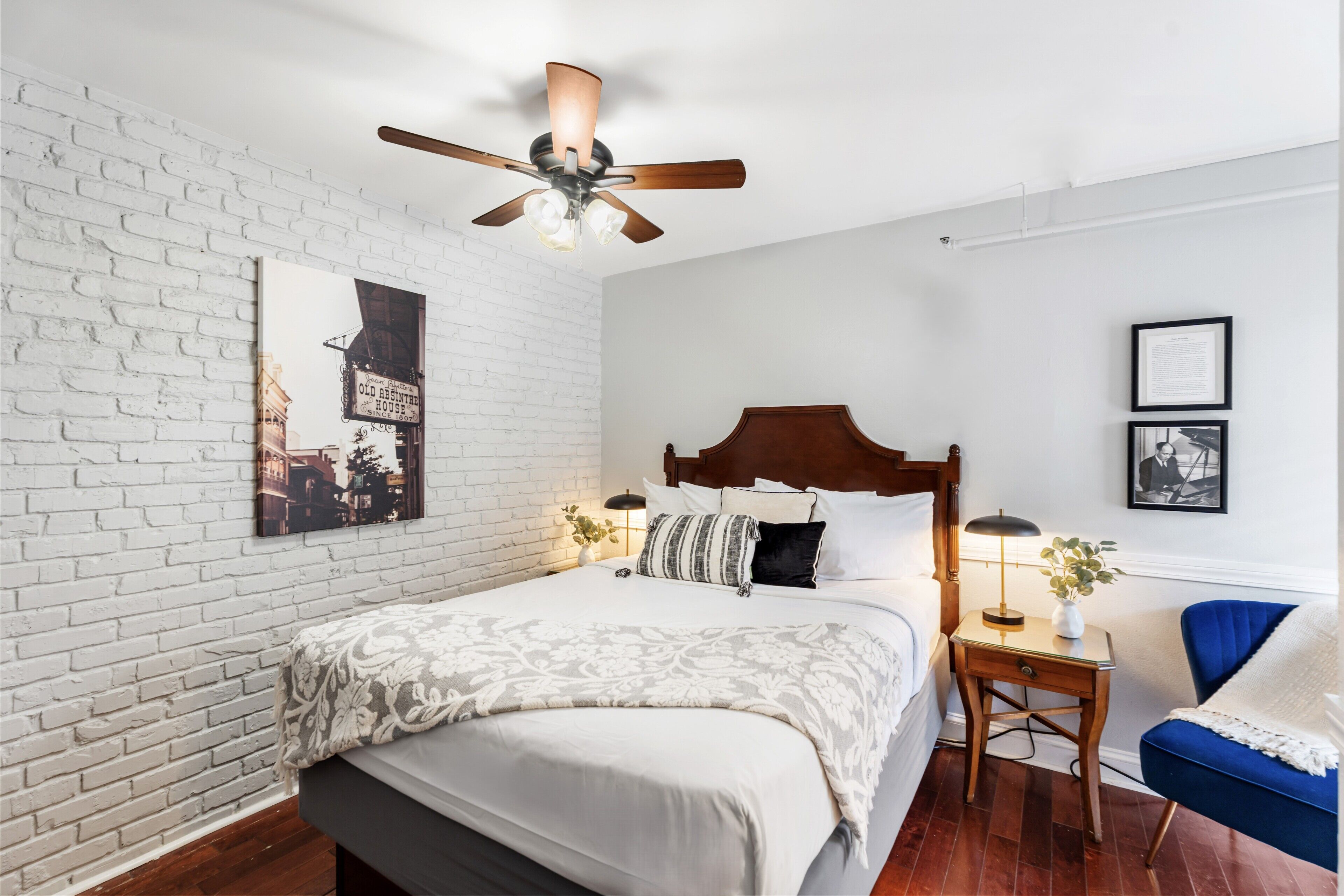 A hotel room with a bed, a nightstand, a chair, a ceiling fan, and a brick wall.