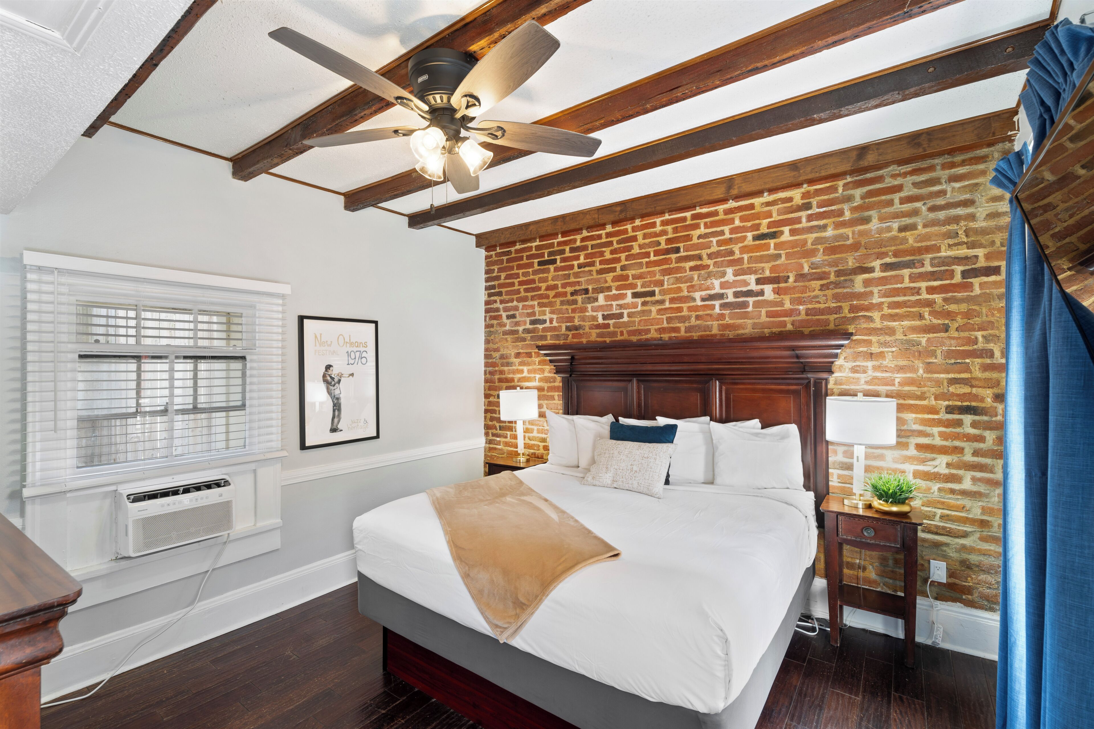 Un dormitorio con pared de ladrillo, ventilador de techo de madera, cama con ropa de cama blanca, mesita de noche con lámpara y ventana con persianas.