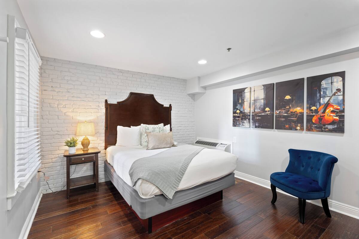 Un dormitorio con cama, mesita de noche, silla, pared de ladrillo y cuadros colgados.