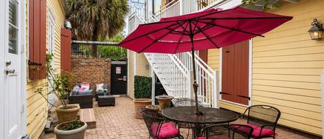 Un patio con una mesa redonda y sillas, una sombrilla roja, y una escalera que lleva a un nivel superior.