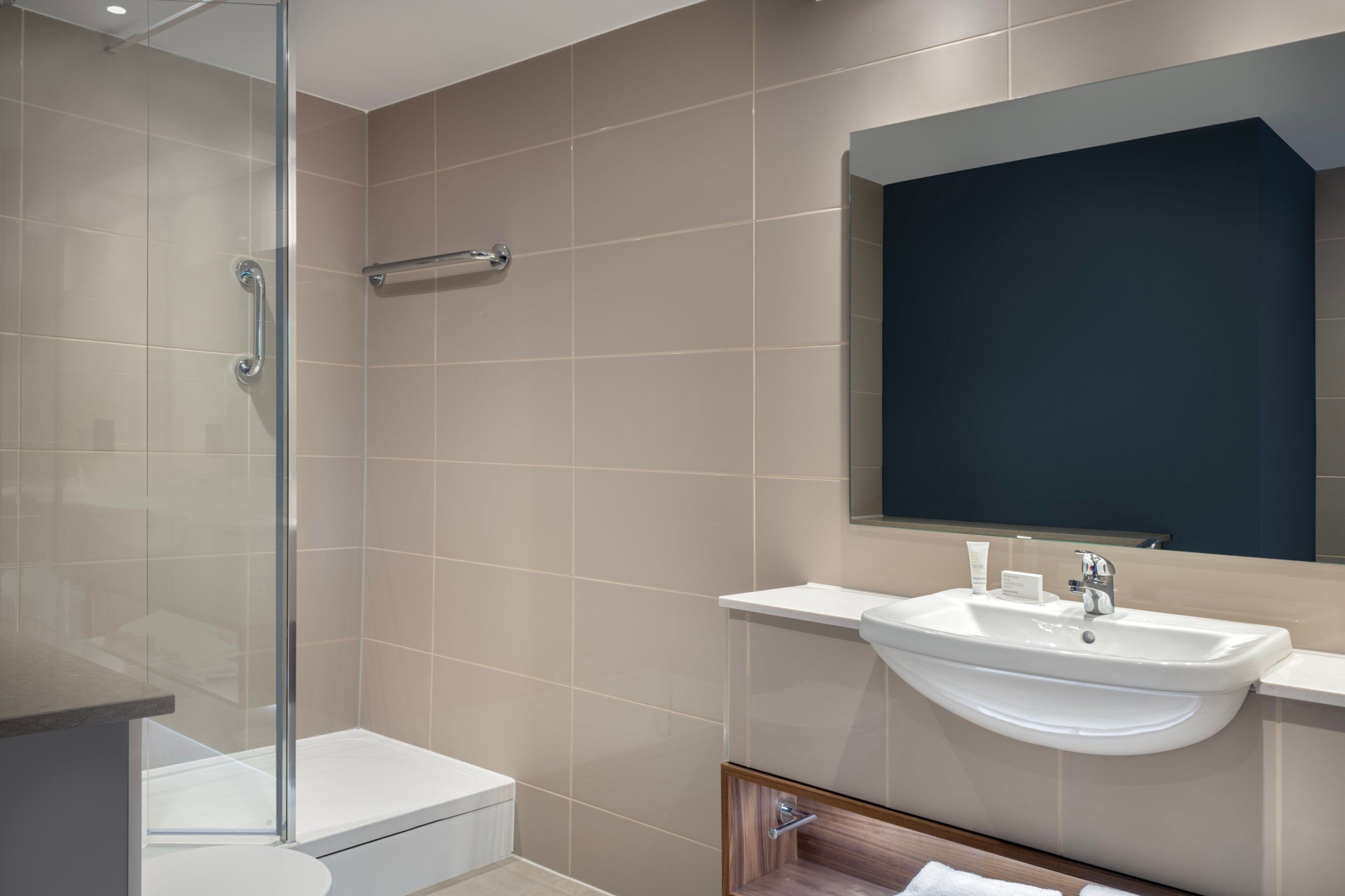 Une salle de bain moderne avec une douche à l’italienne en verre, un lavabo blanc et un miroir aux tons foncés.