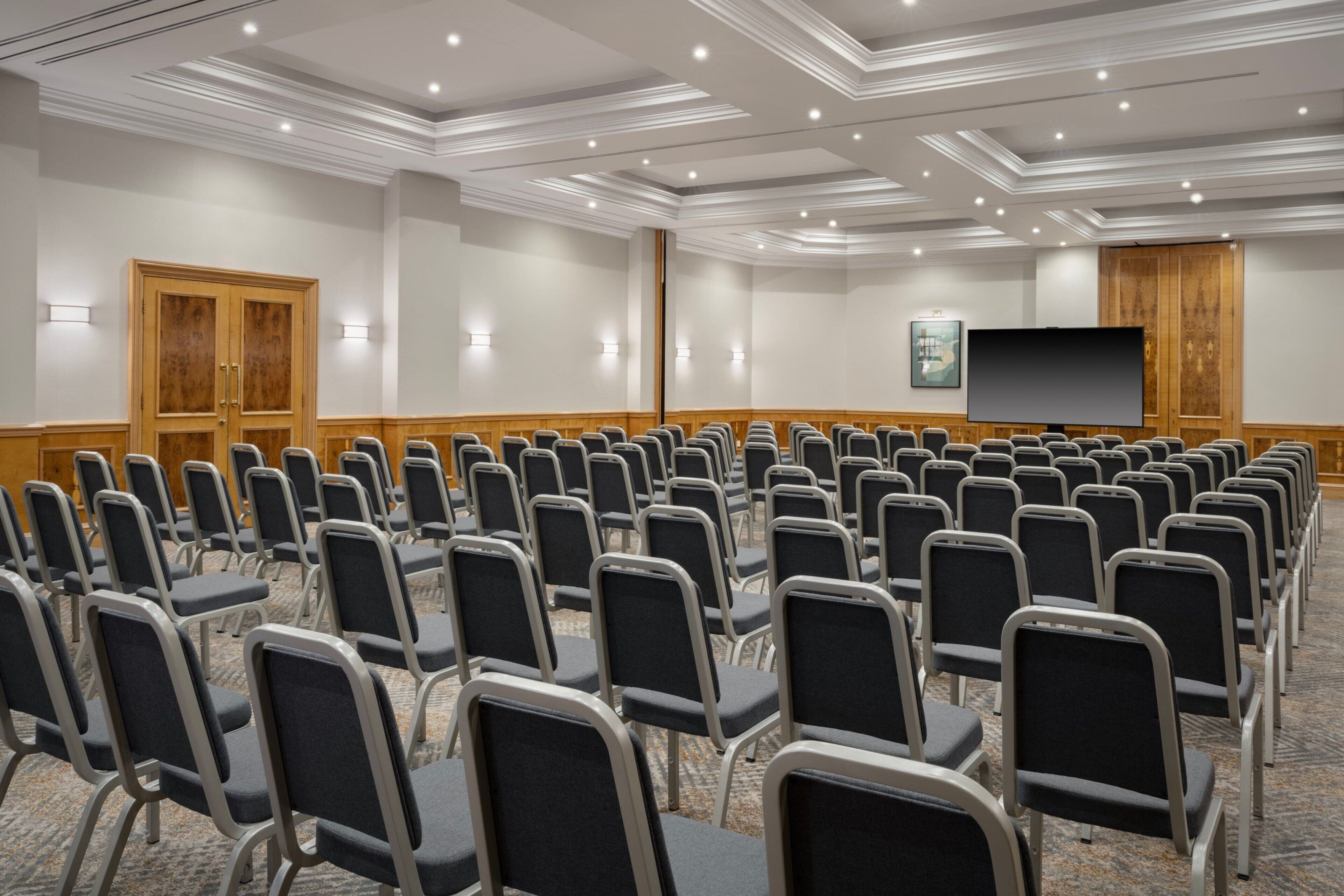 A conference room with rows of chairs, a large screen, and wooden paneling on the walls.